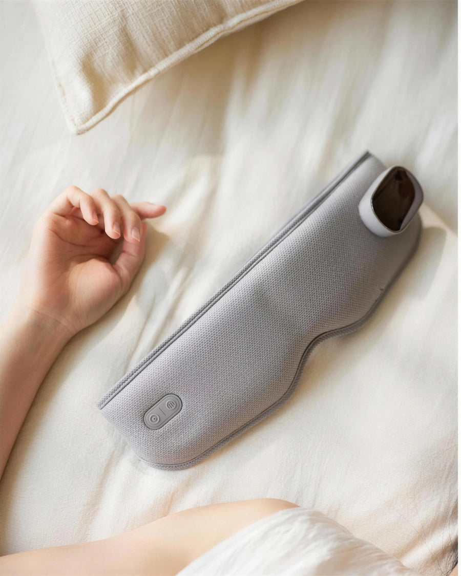 The heated eye massager folded on the bed beside a resting hand