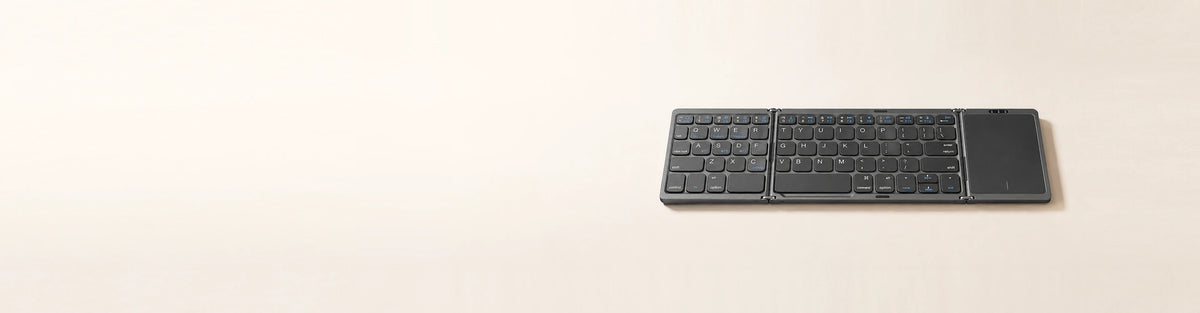 A tri-fold Bluetooth keyboard with a touchpad, unfolded flat on warm oatmeal ground in soft morning light