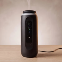Waterless diffuser in Ink Black releasing a thin wisp of oil mist, its cable running off to the right
