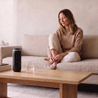 Waterless diffuser in Ink Black on an oak coffee table, a woman resting on the sofa beside it